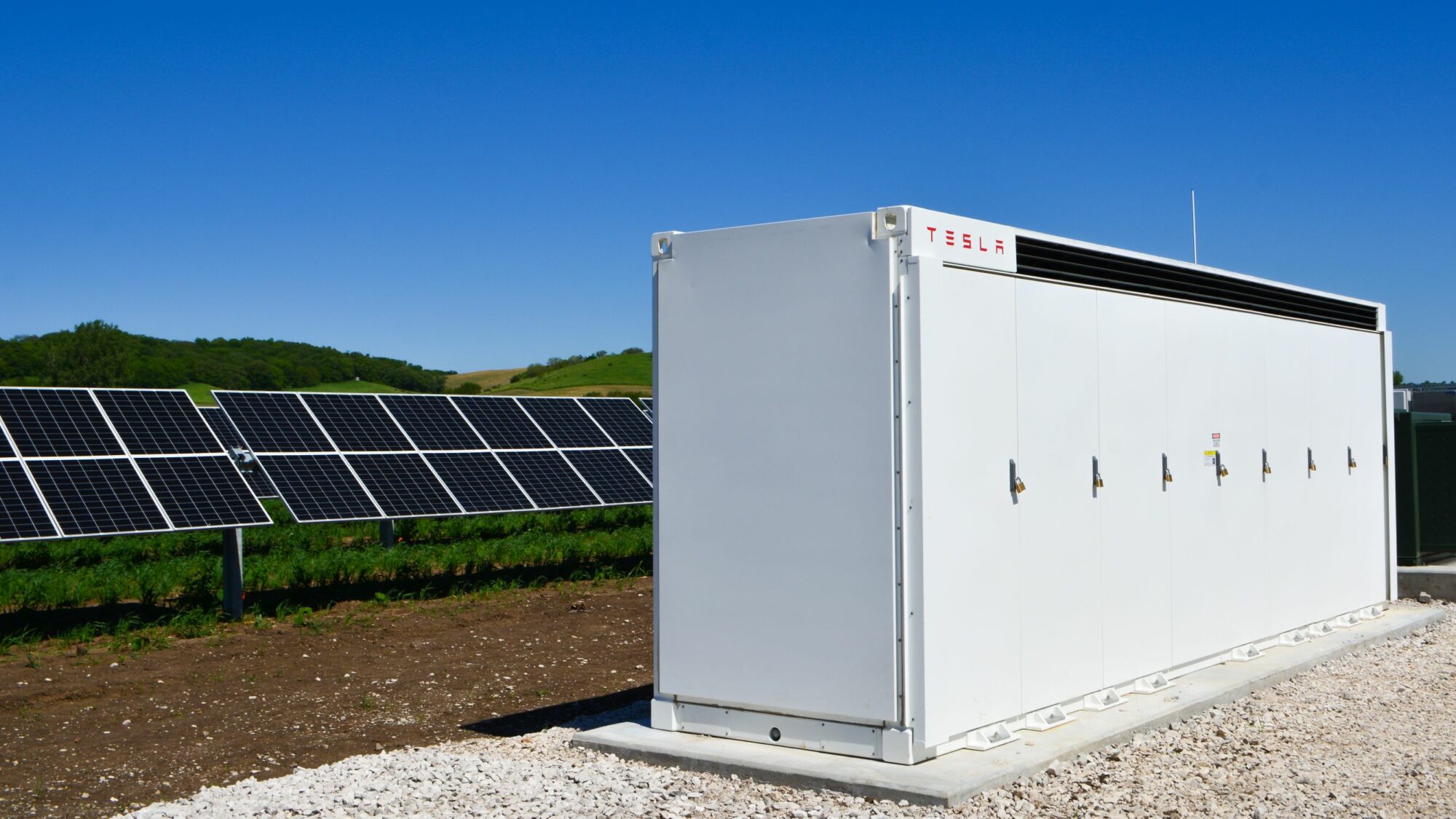 Tesla Megapack with solar panels behind it on an open field with a clear blue sky in the background.